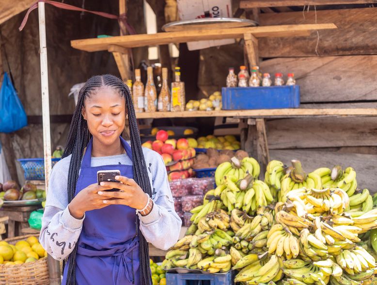 An African woman holding a mobile phone in her hands and looking at the screen while standing in front of a fruit stall in an outdoor market. Image by Wazzkii via Shutterstock.com.