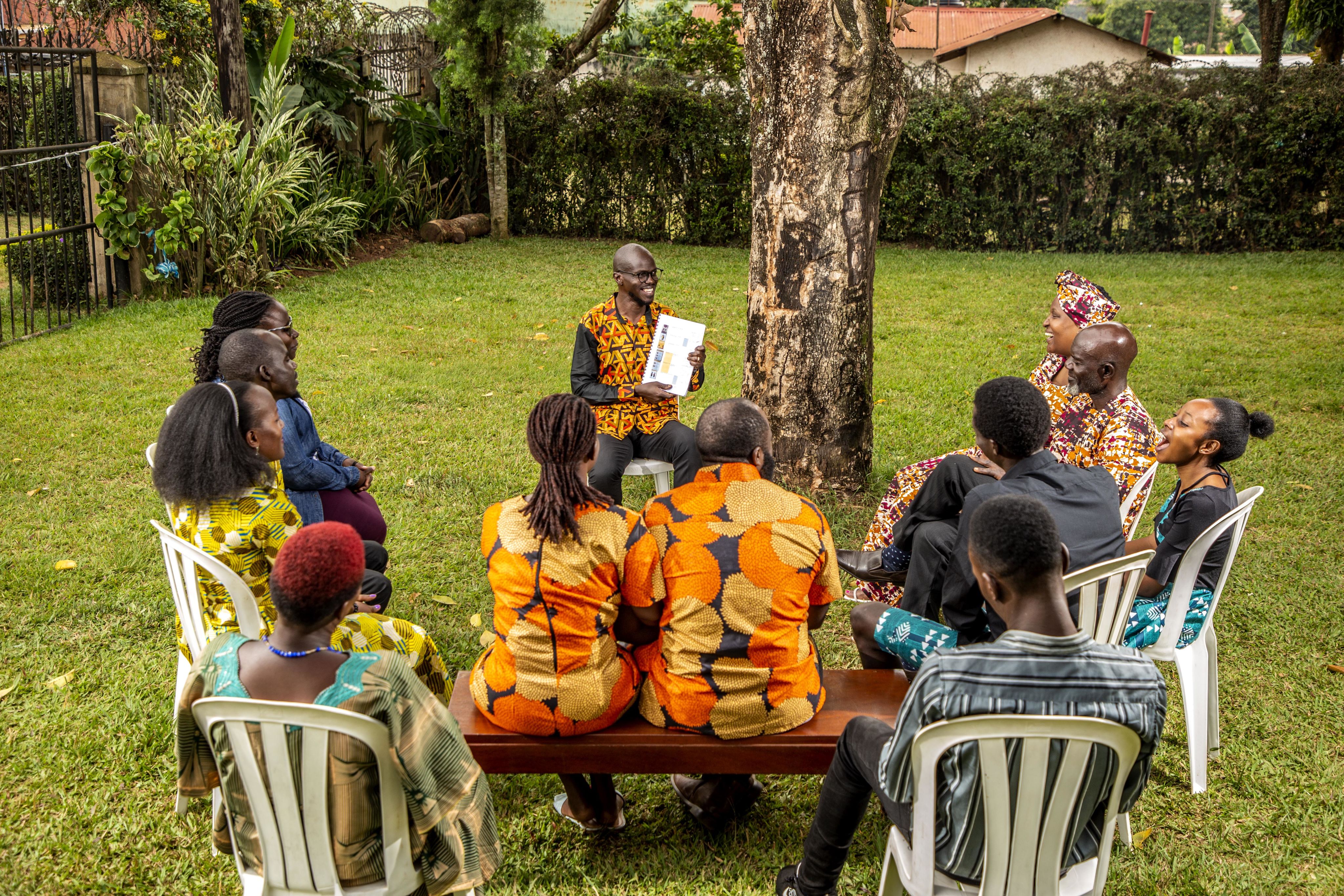 A faith leader holding a piece of paper sits in a chair smiling while facing a group of five couples sitting in chairs in a circle around him, who look happy and excited to be there